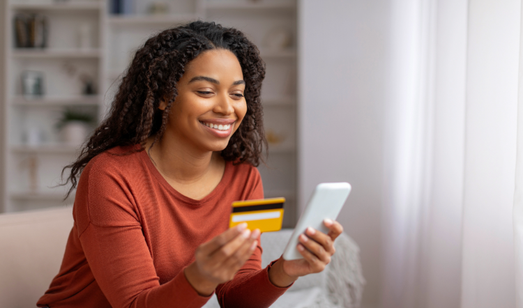 A smiling woman holds a debit or credit card in one hand and a smartphone in the other.